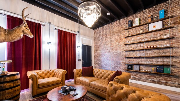 A modern living room with leather sofas, a chandelier, and a brick wall with shelves of books and glasses. - The Rickhouse in NuLu~2 King Bed/2 Full Bath (Louisville)