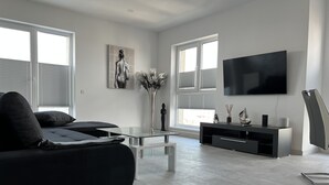 Una sala de estar moderna con un sofá, una mesa de centro de vidrio, un televisor y un ventanal con persianas.