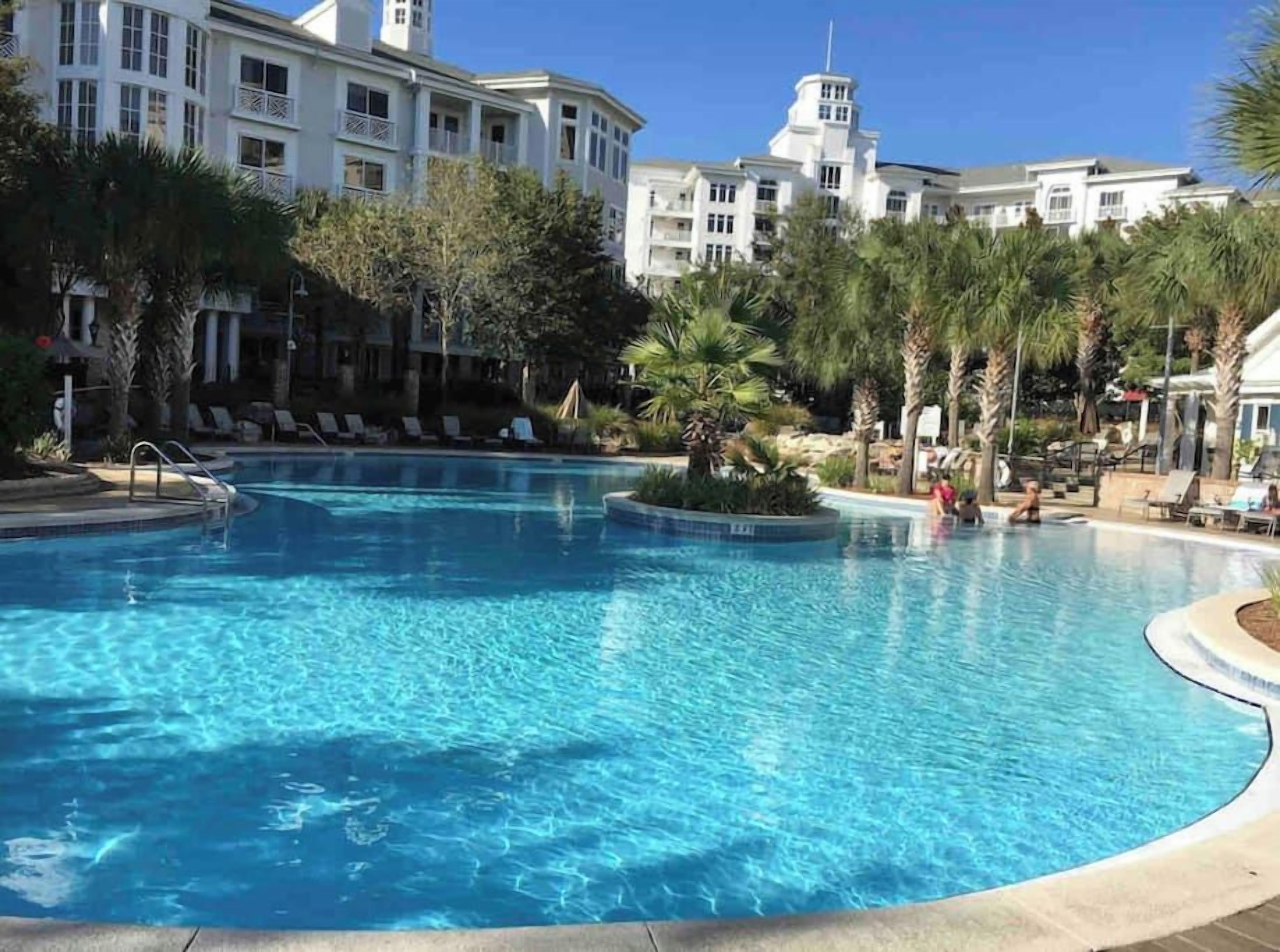 A swimming pool with lounge chairs and palm trees, surrounded by a building with multiple balconies.