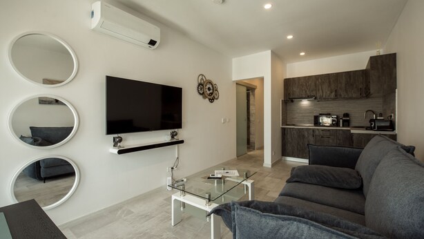 Ein modernes Wohnzimmer mit Flachbildfernseher, drei Wandspiegeln, einem Glastisch und einem grauen Sofa.