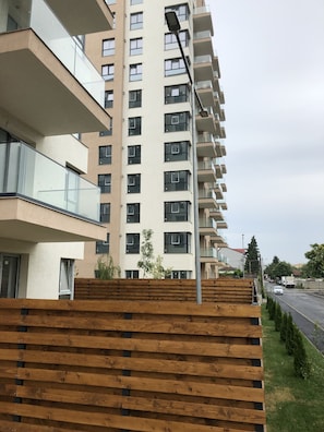 A multi-story residential building with balconies, a street in the background, and a wooden fence in the foreground.