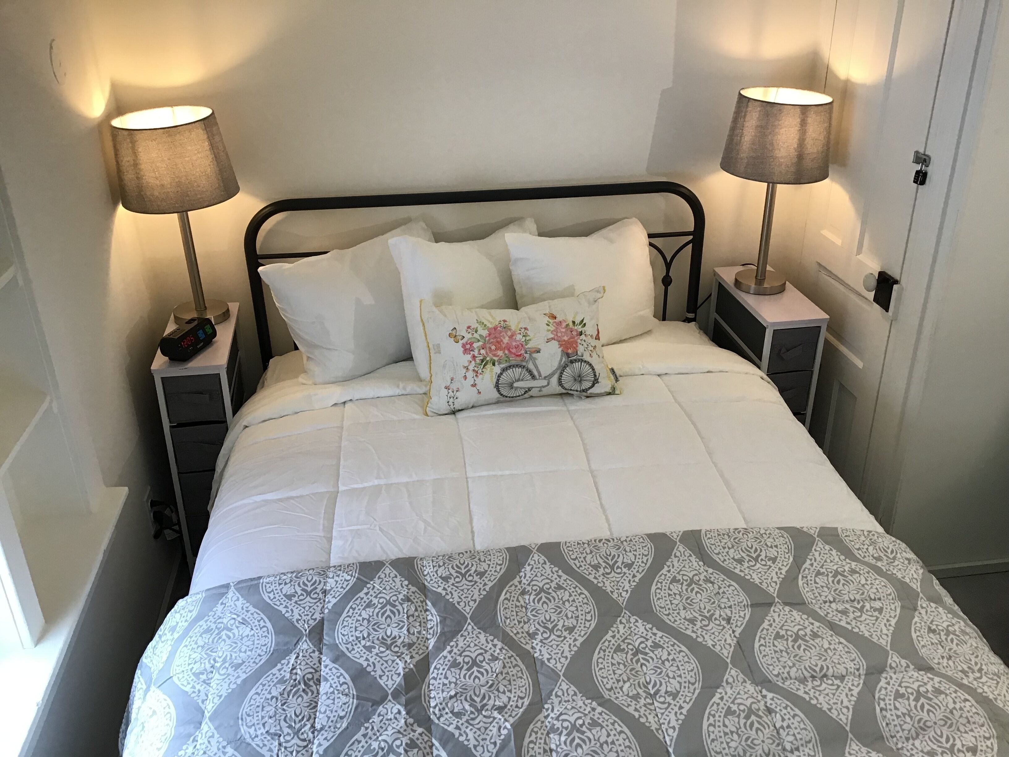 Cama bem arrumada com colcha estampada, duas luminárias de cabeceira, um relógio e um travesseiro decorativo.