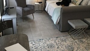 Una habitación acogedora con una cama, un escritorio, una silla y una mesita pequeña. Hay dos ventanas con persianas, un ventilador de techo y una alfombra estampada.