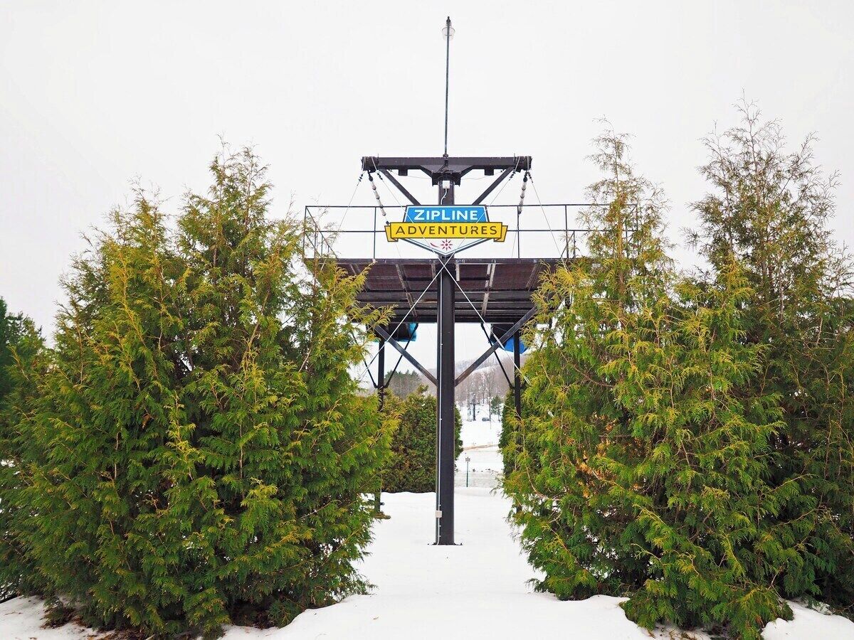 Segnaletica per Zipline Adventures, con struttura metallica e sentiero innevato affiancato da alberi.
