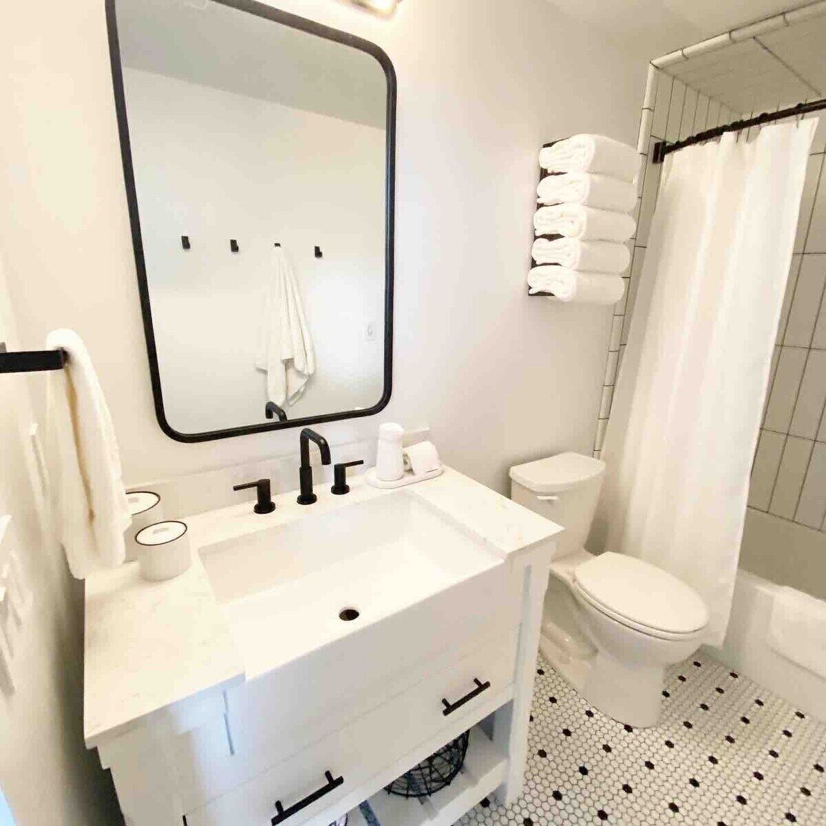 Un baño con un lavamanos blanco, espejo y retrete. Hay toallas apiladas en la pared y una cortina de ducha.