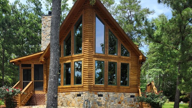 Una cabaña de madera con base de piedra, ventanas grandes y un patio con mobiliario exterior.
