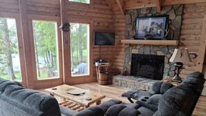 Interior acogedor de una cabaña con chimenea de piedra, un televisor de pantalla plana y un amplio ventanal con vista a un bosque.