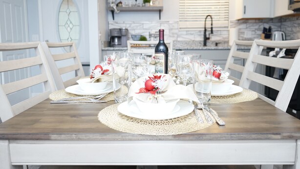 Een eettafel gedekt voor vier personen, met witte borden, helder glaswerk en een fles wijn. De tafel staat in een keuken met witte keukenkastjes en een raam met jaloezieën.