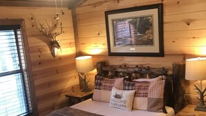Ein gemütliches Schlafzimmer im Blockhausstil mit Holzboden, einem Bett mit Karo-Bettwäsche, einem gerahmten Bild über dem Bett und einem Deckenventilator.