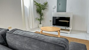Un salon avec un canapé gris, une petite table en bois, un grand téléviseur, une cheminée blanche et une plante en pot.