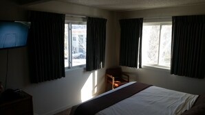 Une chambre d’hôtel avec un lit, une chaise, une télévision et deux fenêtres avec des rideaux.