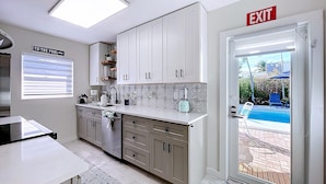 Een moderne keuken met witte kastjes, een ingebouwde oven en een aanrecht met een blender. Een bordje wijst de uitgang aan en er is een uitzicht op een zwembad buiten.