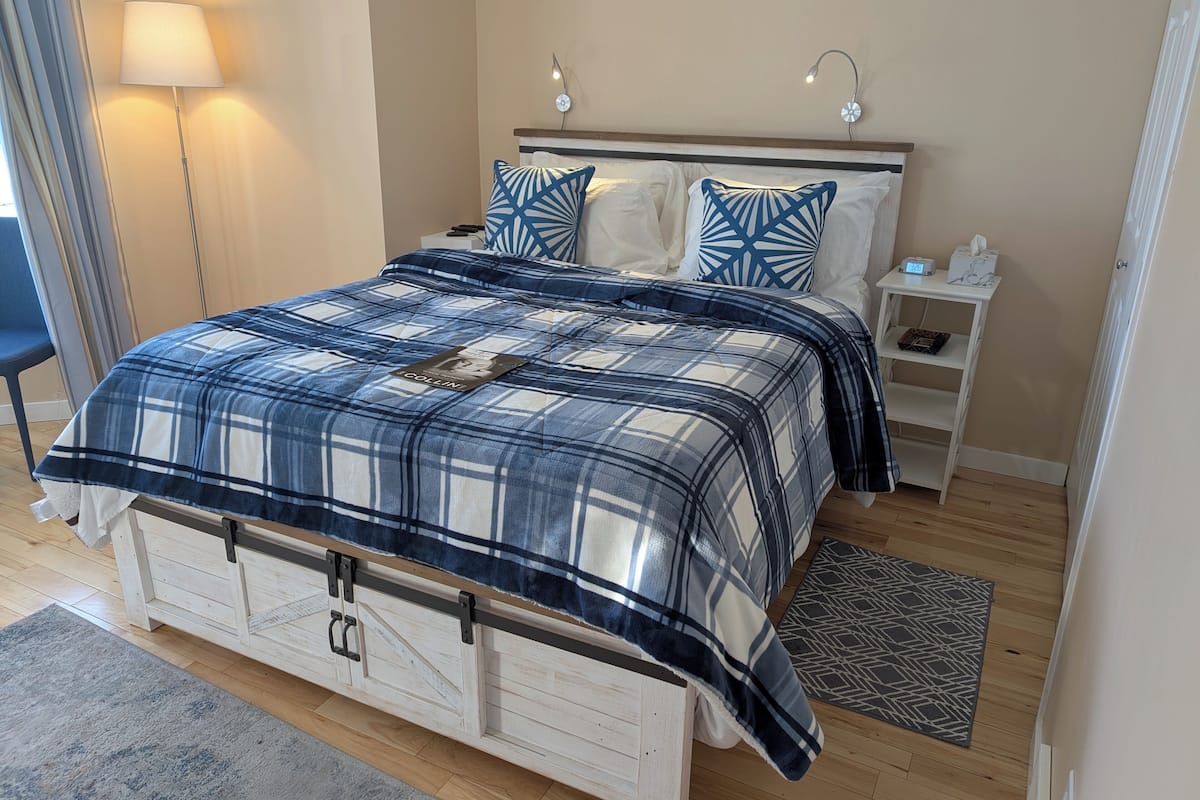 A bedroom with a plaid bedspread, a bedside table with a lamp, and a nightstand with a phone.