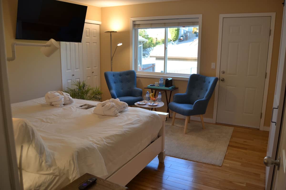 A hotel room with a bed, two armchairs, a small table with drinks, a TV mounted on the wall, and a window with a view of the outside.
