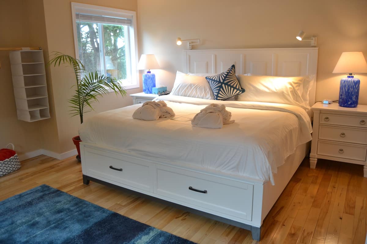 A neatly made bed with two towels, a bedside table with a lamp, a nightstand with a lamp, a window with blinds, and a plant.