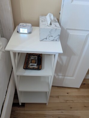 Una pequeña mesita de noche blanca con un reloj digital, una caja de pañuelos y una bandeja decorativa.