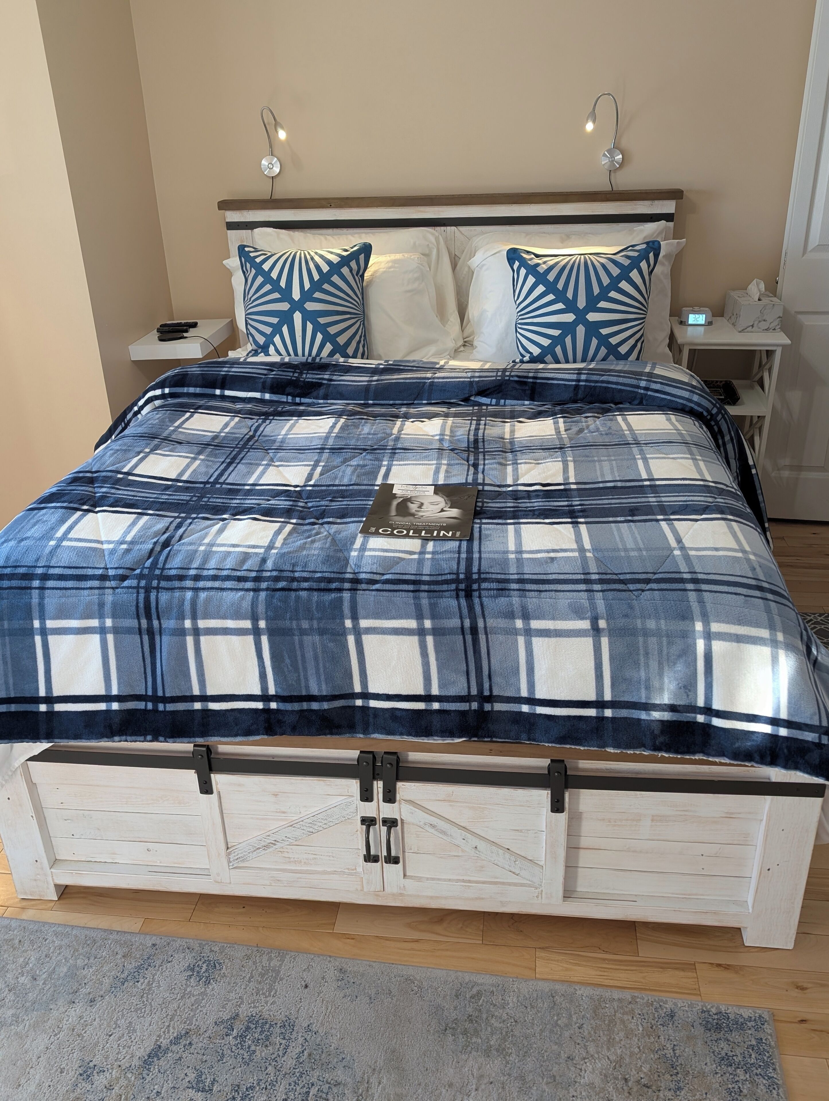 Una cama con una colcha escocesa, dos almohadas con diseño geométrico, una mesita de noche con un reloj y un mueble auxiliar con una caja de pañuelos.