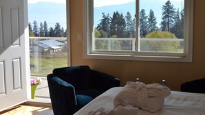 Una habitación con cama, ventana con vista a árboles y montañas, una silla y un florero con flores.