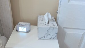 Una mesita de noche blanca con un reloj digital, una caja de pañuelos y una bandeja decorativa.