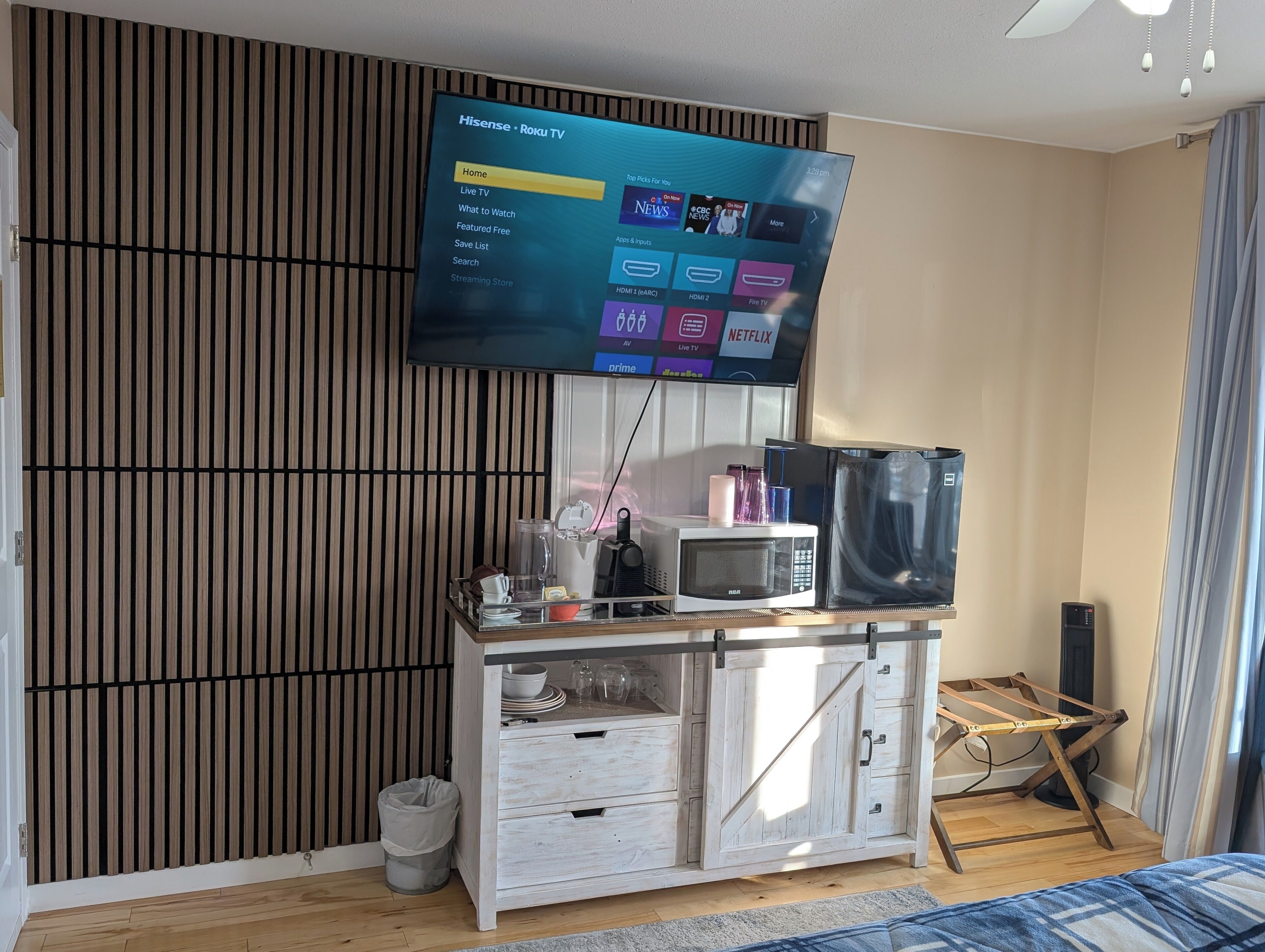 A modern living room with a mounted TV displaying a Roku interface, a wooden cabinet with drawers, a microwave, and a small trash bin.
