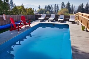 Una piscina rectangular con un deck de madera, rodeada de sillas de lounge blancas y una silla roja.