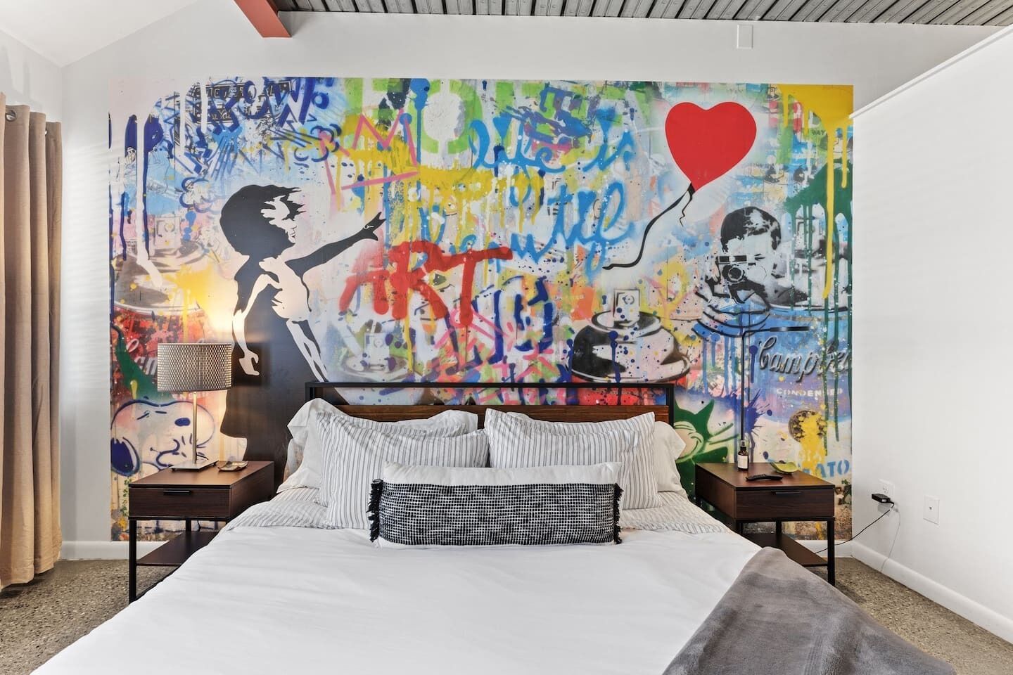 Une chambre à coucher avec un grand lit, une tête de lit ornée d’un motif en forme de cœur et une fresque colorée sur le mur.