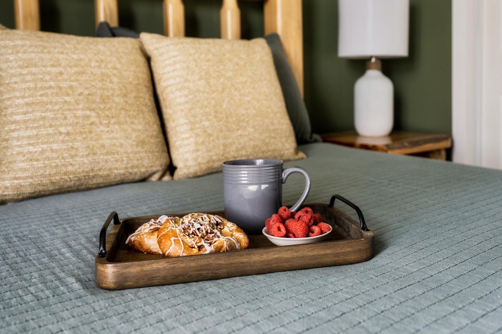 Una cama con un edredón gris, una bandeja de madera con un pastel, frambuesas y una taza gris.