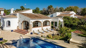 Blick auf einen Pool vor einer Villa im mediterranen Stil, mit Sitzgelegenheiten im Freien und üppiger Bepflanzung.