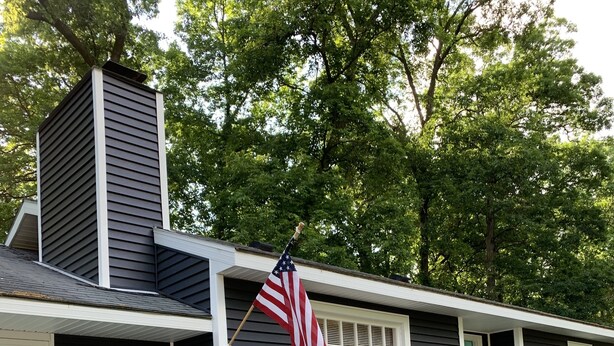 Ein Haus mit dunkler Fassade, einem Schornstein, einer Veranda mit zwei Stühlen, einer amerikanischen Flagge und einem Briefkasten.