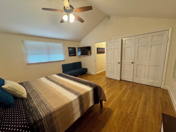 A bedroom with a bed, a ceiling fan, a window with blinds, a sofa, and a set of white doors.