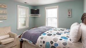 Ein Schlafzimmer mit einem Bett, zwei Fenstern mit Jalousien, einem Fernseher auf einem Regal und einem Sessel.