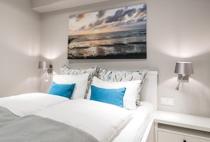 A modern bedroom with a large bed, two wall-mounted lamps, and a painting of a sunset over the ocean. - Loddis Hüs - Loddi's Hüs (Wenningstedt-Braderup (Sylt))