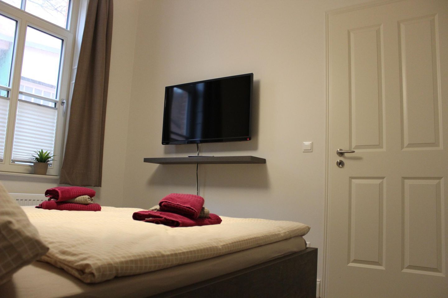 Ein Schlafzimmer mit einem Bett, einem an der Wand befestigten Fernseher, einer Tür und einem Fenster mit Jalousien.