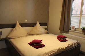 Una cama bien hecha con dos almohadas y dos toallas dobladas, una lámpara de noche y una ventana con persianas.
