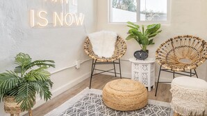 Ein gemütliches Zimmer mit einem großen Fenster, Korbstühlen, einem Rattan-Hocker und einem Wandleuchtschild mit der Aufschrift 'ALL WE HAVE IS NOW'.