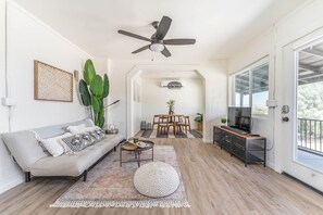 Ein geräumiges Wohnzimmer mit einer Couch, einem Couchtisch und einem Essbereich mit Stühlen. Ein Deckenventilator und ein Fernseher sind zu sehen.