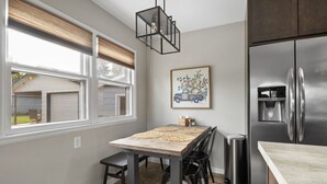 A modern kitchen with a stainless steel refrigerator, a dining table with black chairs, and a window with blinds.