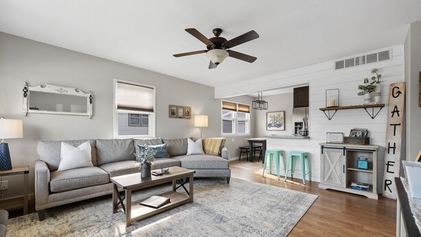 A living room with a grey sofa, a coffee table, and a ceiling fan. There is a kitchen area with a bar counter and stools.