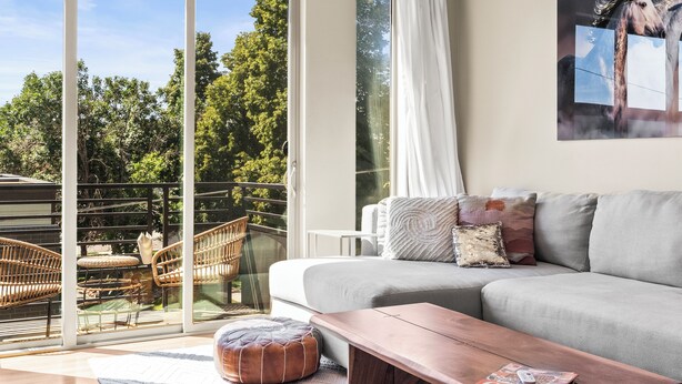 Un salon moderne avec un canapé gris, une table basse en bois et une grande porte-fenêtre coulissante donnant sur un balcon extérieur aménagé avec des meubles en osier.
