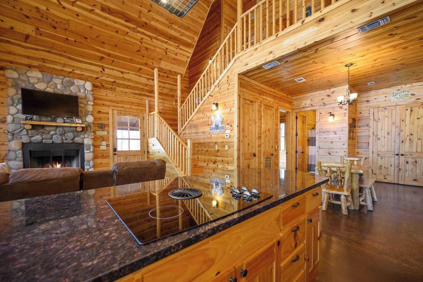 A log cabin interior with a stone fireplace, a wooden staircase, a dining area, and a kitchen with wooden cabinets.