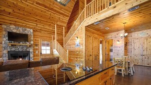 Interior de uma cabana de tronco com lareira de pedra, escada de madeira, área de jantar e cozinha com armários de madeira.