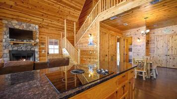 A log cabin interior with a stone fireplace, a wooden staircase, a dining area, and a kitchen with wooden cabinets.