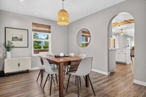 Un espace repas moderne avec une table en bois, des chaises blanches, un luminaire suspendu et une vue sur la cuisine, accessible par une arche.