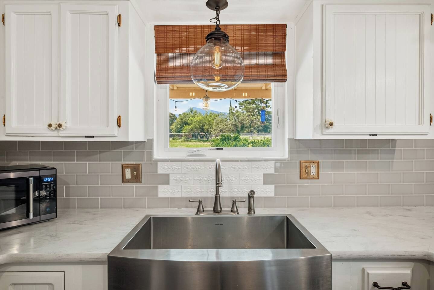 A kitchen with white cabinets, a stainless steel sink, a microwave, and a window with a view of greenery.