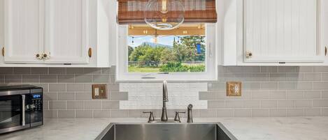 A kitchen with white cabinets, a stainless steel sink, a microwave, and a window with a view of greenery.