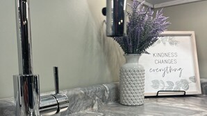 Un lavamanos de cocina con grifo cromado, encimera de mármol, un florero blanco con flores moradas y una cita enmarcada.