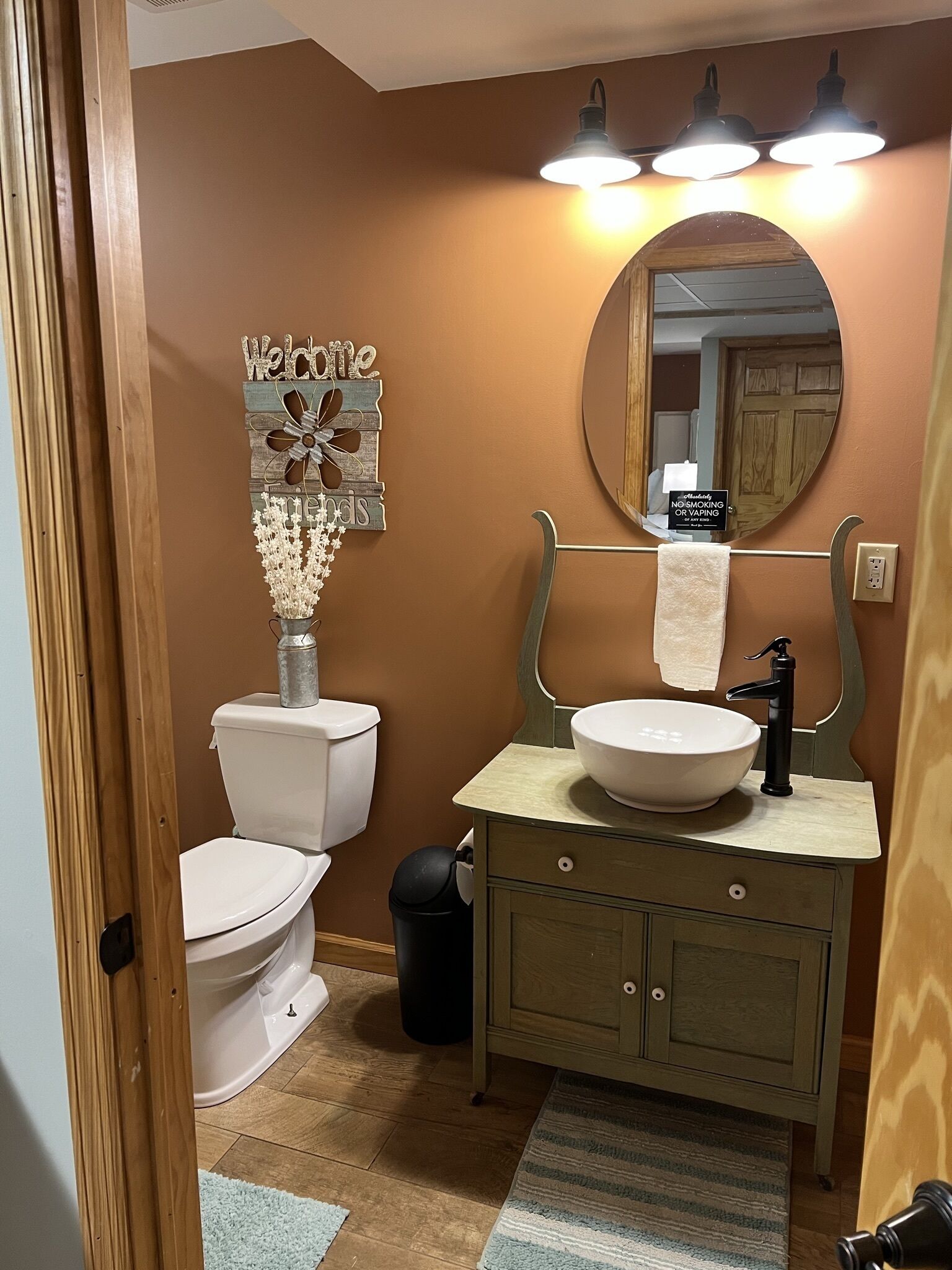 A bathroom with a toilet, sink, and mirror. There is a 'Welcome' sign and a 'No Smoking' sign on the wall.