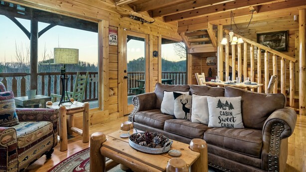 A cozy cabin interior with a brown leather couch, wooden furniture, and a scenic view through large windows. - SPRING DISCOUNTS! Hot Tub ⭐️ AMAZING VIEWS⭐️ Games 🎱 🐻 (Sevierville)