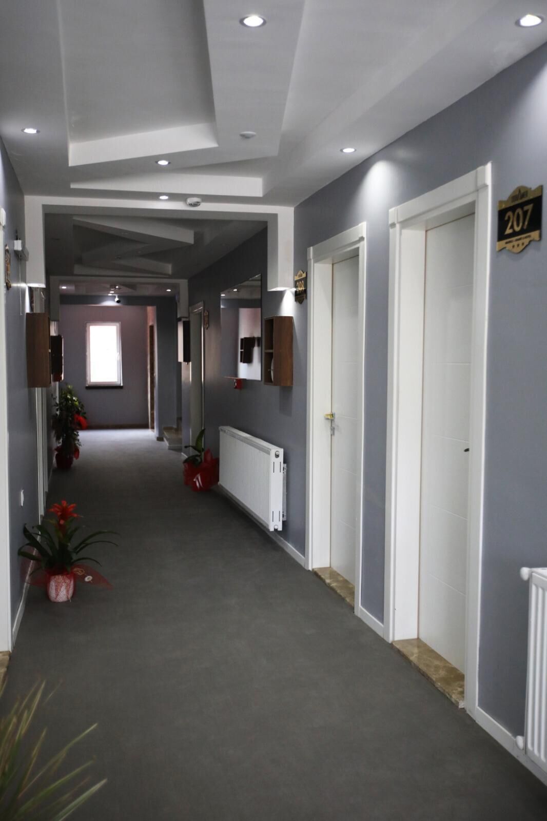 A hallway with grey walls, white doors, and a radiator. There are potted plants along the left side and a number sign on the right wall.