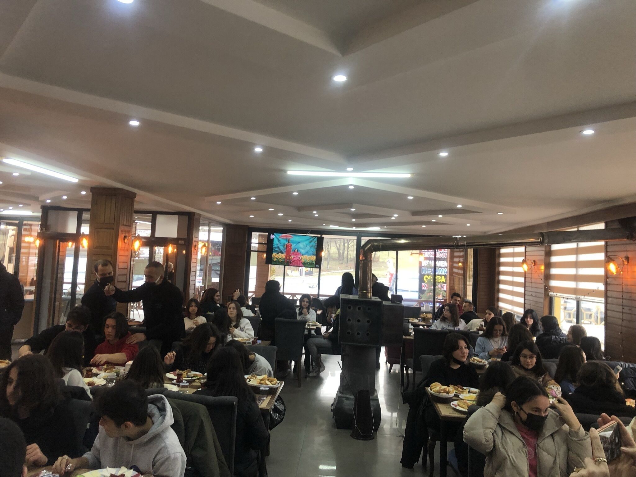 A restaurant with multiple tables and chairs, a television screen, and a large window.